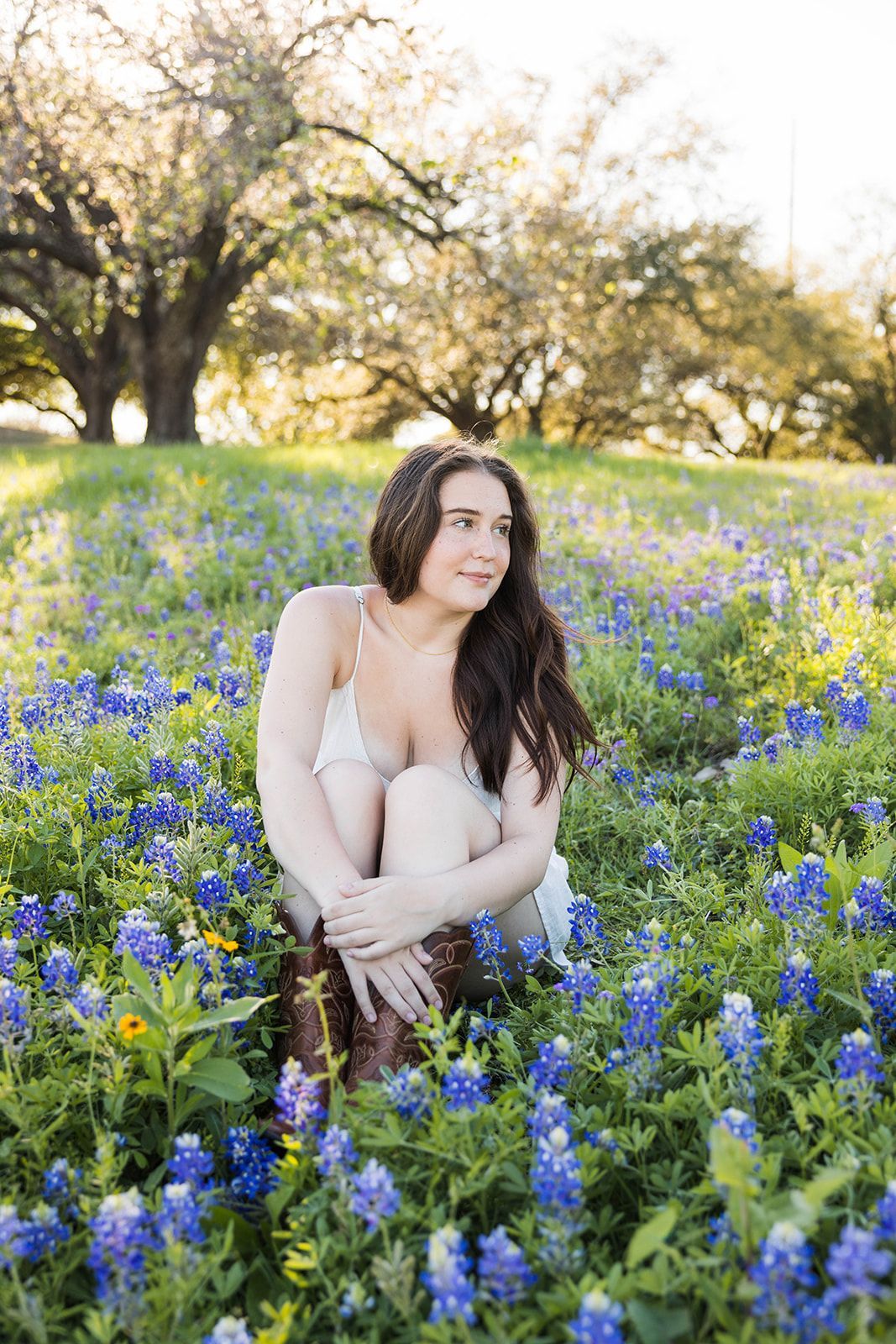 A person sitting in a field of bluebonnets under trees, looking off to the side with a calm expression.