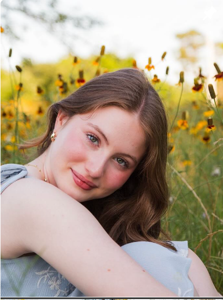 A person with light brown hair leans toward the camera, smiling softly in a field of yellow wildflowers during sunset.