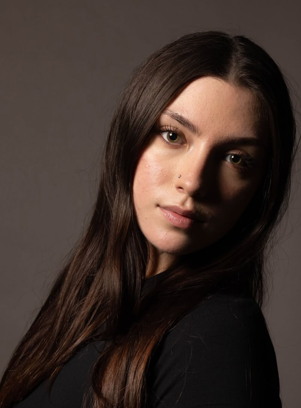 A person with long brown hair posing against a neutral dark background with dramatic, side-lit studio lighting.