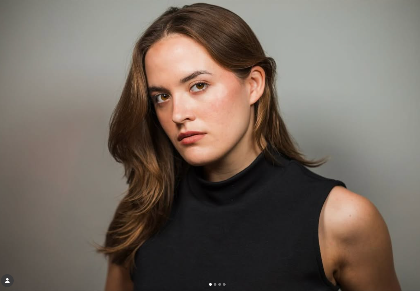 A person with long brown hair wearing a black sleeveless top, looking toward the camera against a gray background.