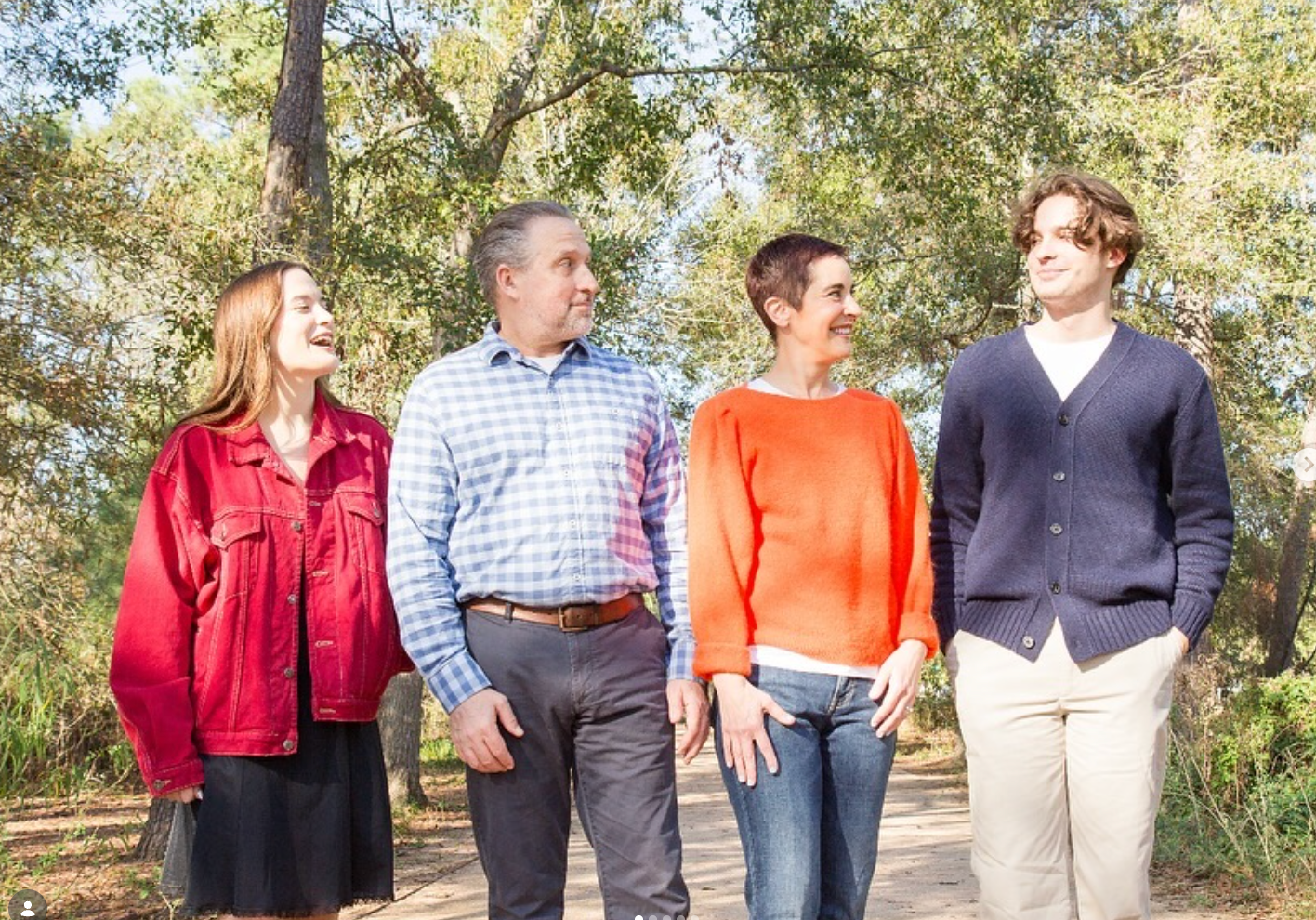 Four people walk along a sunny, tree-lined path, laughing and looking toward each other.