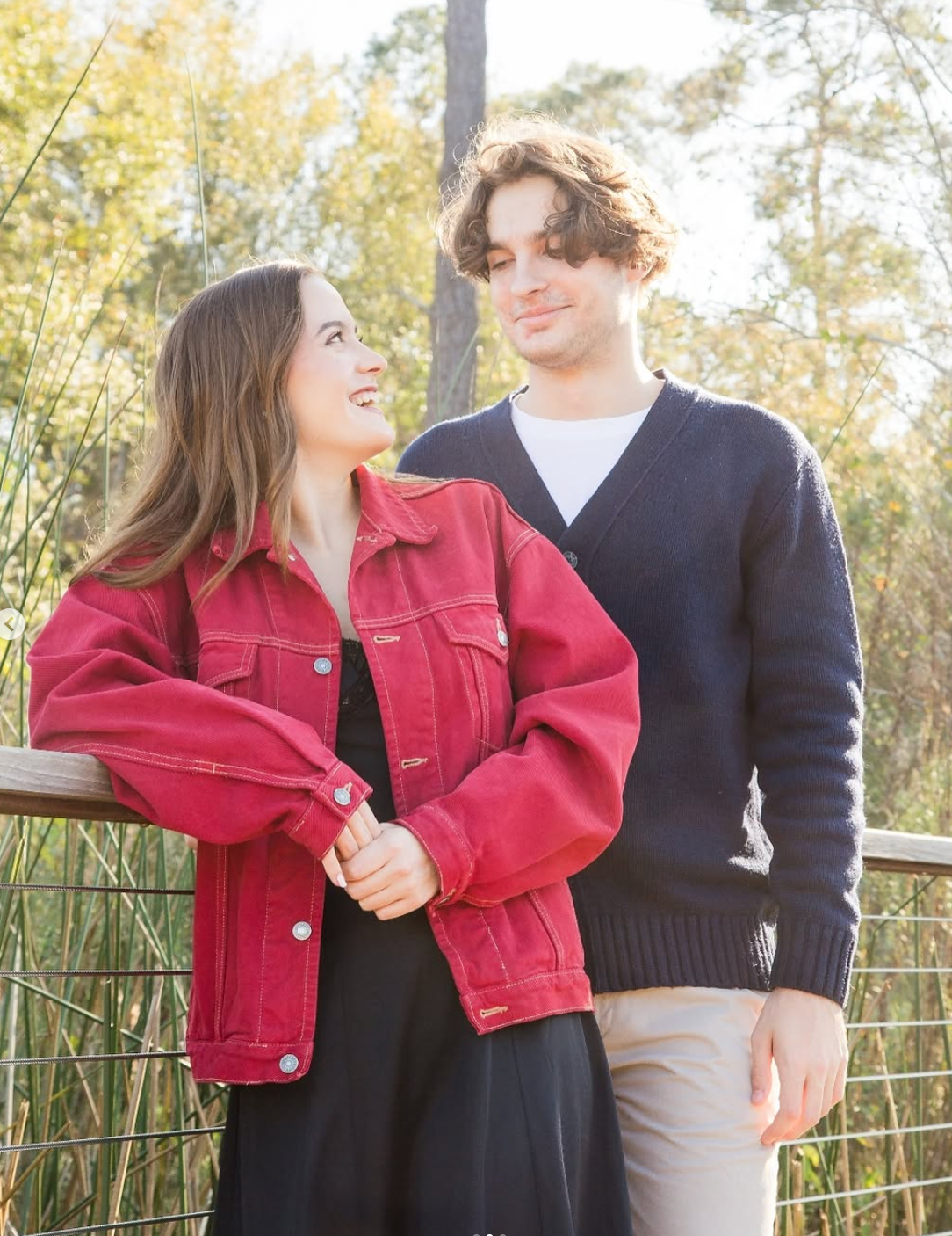A young couple stands smiling at each other on a wooden bridge, wearing a red jacket and a navy blue cardigan.