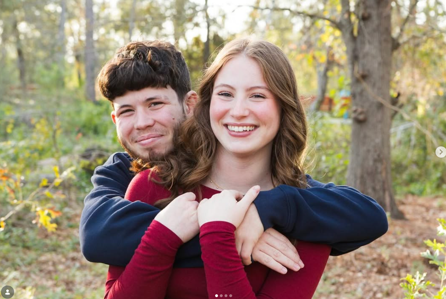 A person in a maroon shirt is smiling while being hugged from behind by a person in a dark blue sweater in a wooded area.