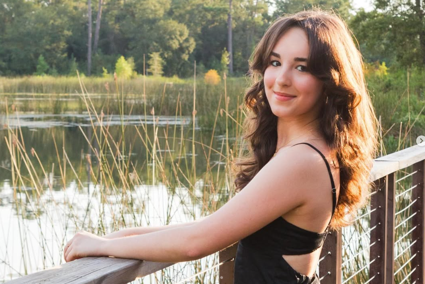 A smiling person with wavy brown hair wearing a black top, leaning on a wooden railing beside a pond and tall grasses.