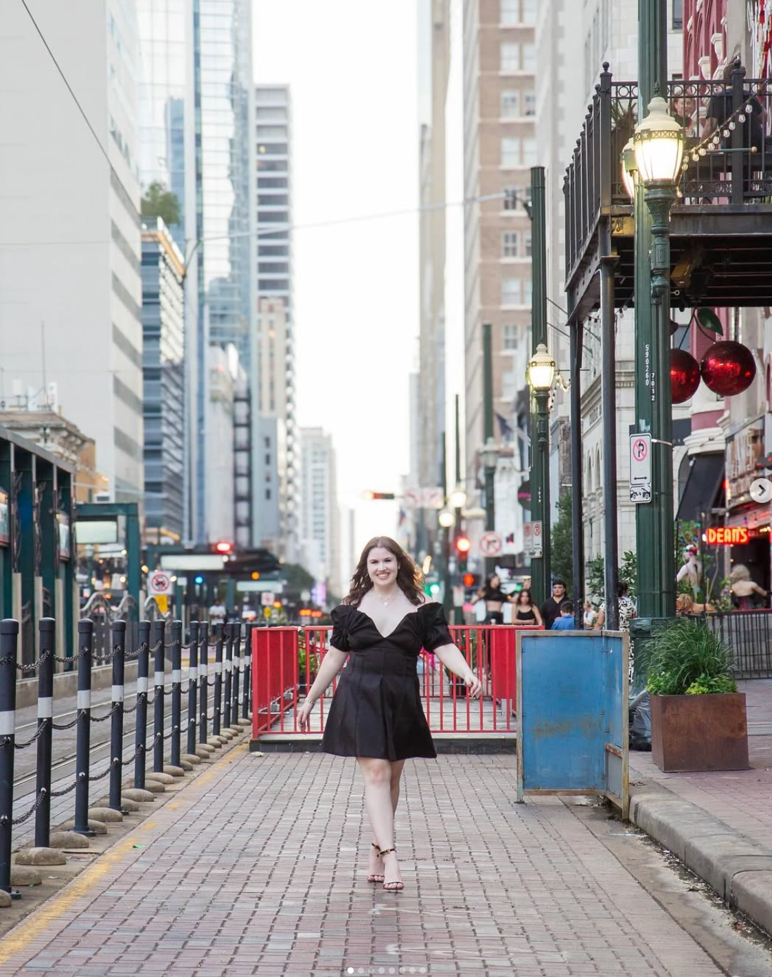A smiling person in a black dress walking toward the camera down a brick sidewalk in a city lined with tall buildings.