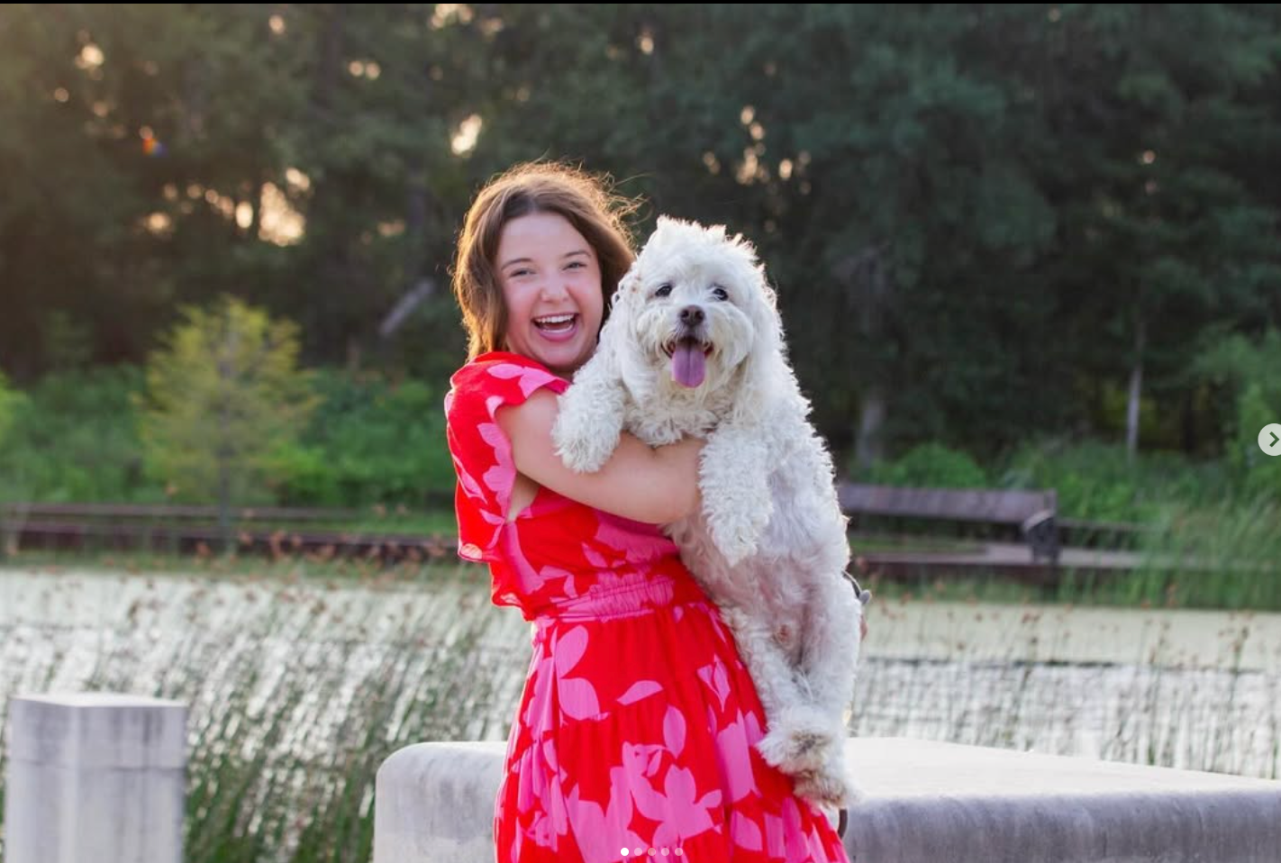 A smiling person in a bright pink floral dress holds a fluffy white dog outdoors near a pond.