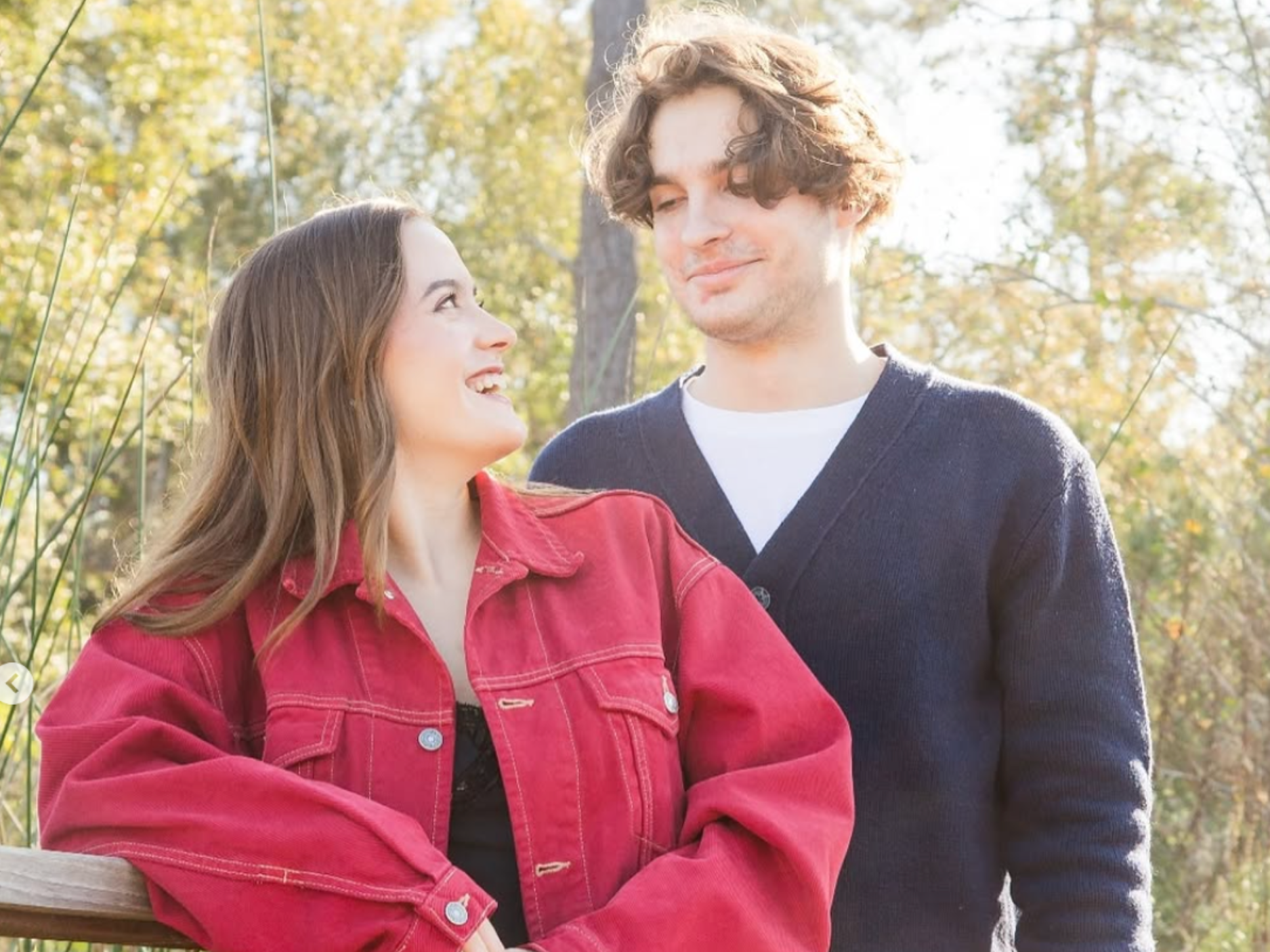 A young woman in a red denim jacket smiles at a young man in a dark cardigan, standing together in a wooded area.