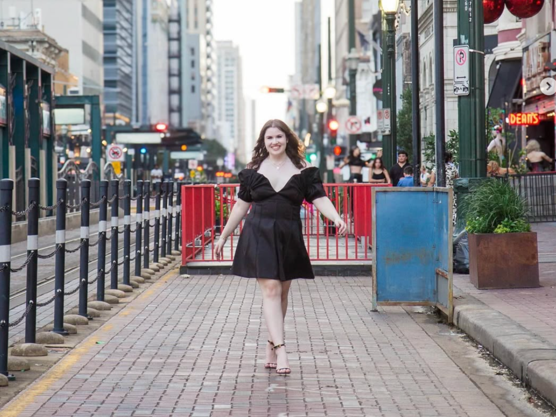 A smiling person in a black dress walks along a paved urban street with tall city buildings and red railings.