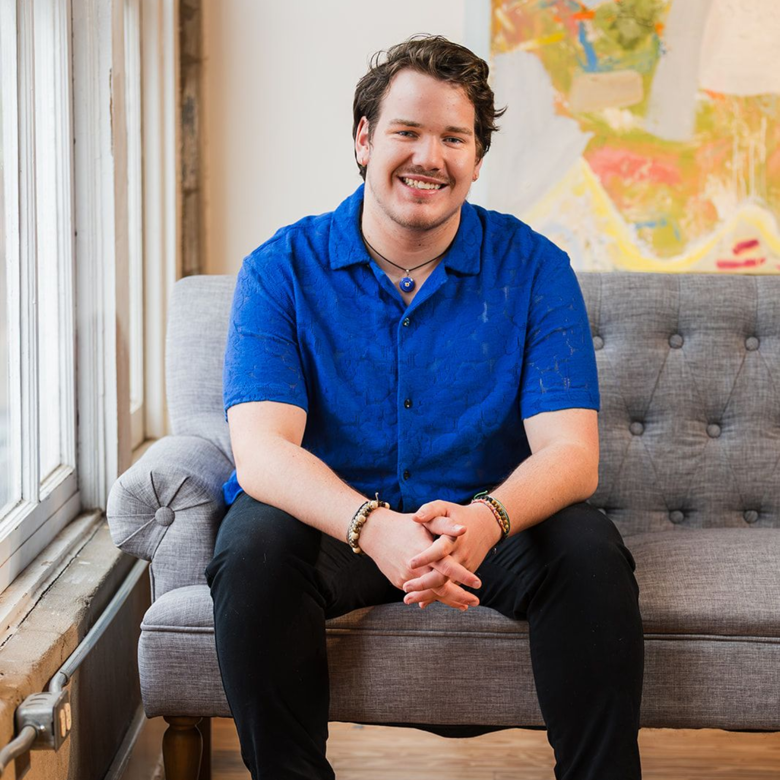 A person wearing a blue collared shirt and bracelets smiles while sitting on a gray tufted couch in a sunlit room.