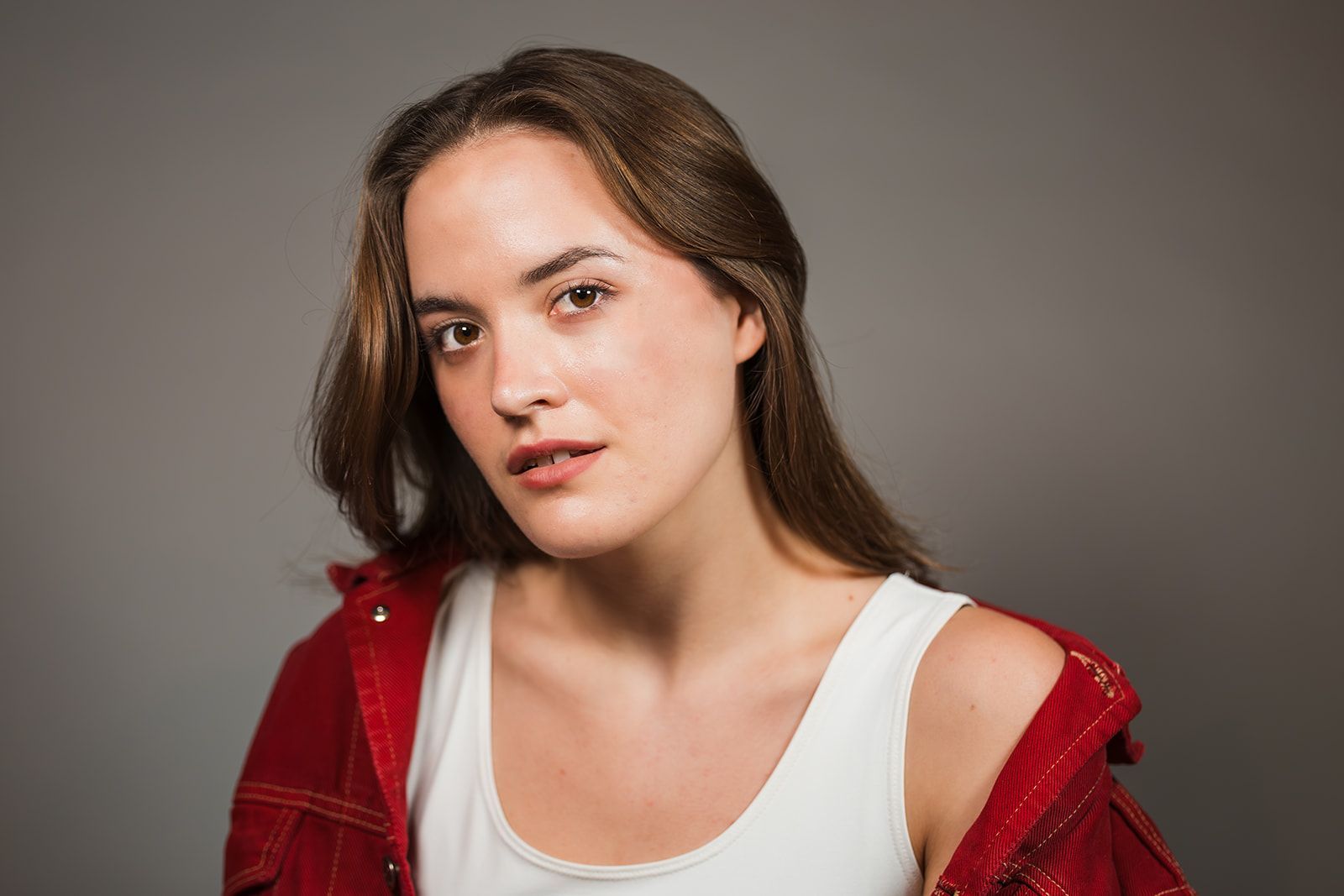 A person with long brown hair, wearing a white tank top and a red jacket, poses against a plain gray background.
