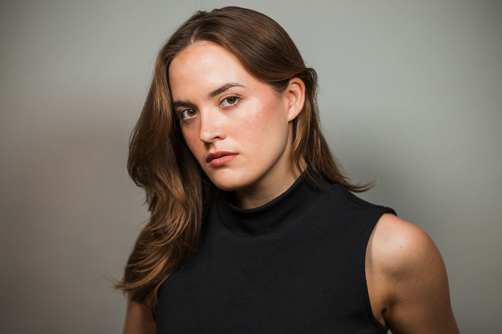 A woman with long brown hair wearing a black sleeveless top, looking toward the camera against a grey background.
