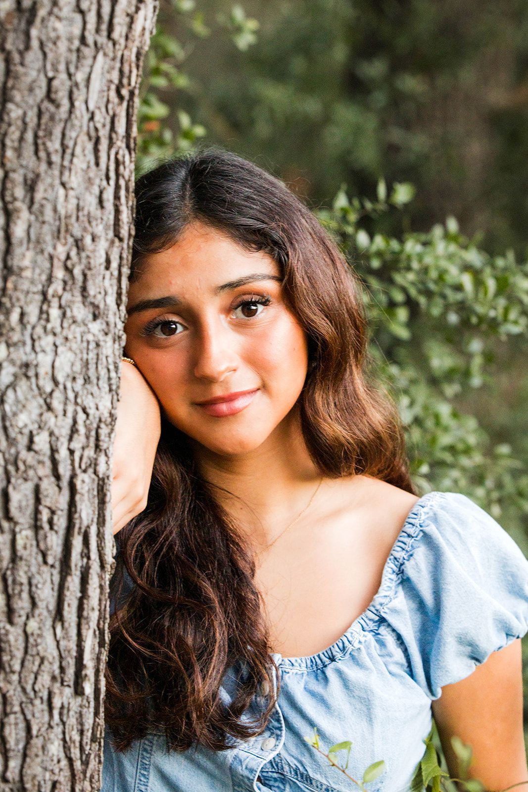 A young person with long, wavy dark hair leaning against a tree trunk in a park setting, looking toward the camera.