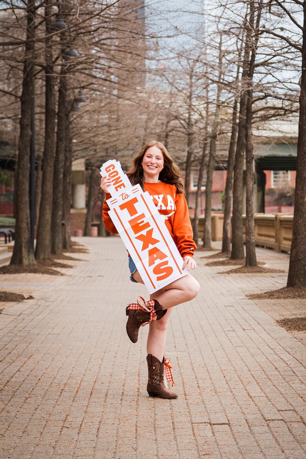 A person stands on a brick path between trees, smiling while holding a "Gone to Texas" sign and wearing cowboy boots.
