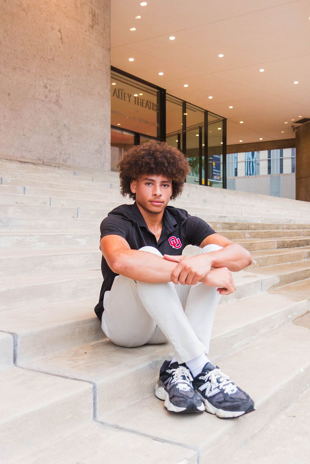 A person with curly hair sits on concrete steps in front of a building, wearing a black polo shirt, pants, and sneakers.
