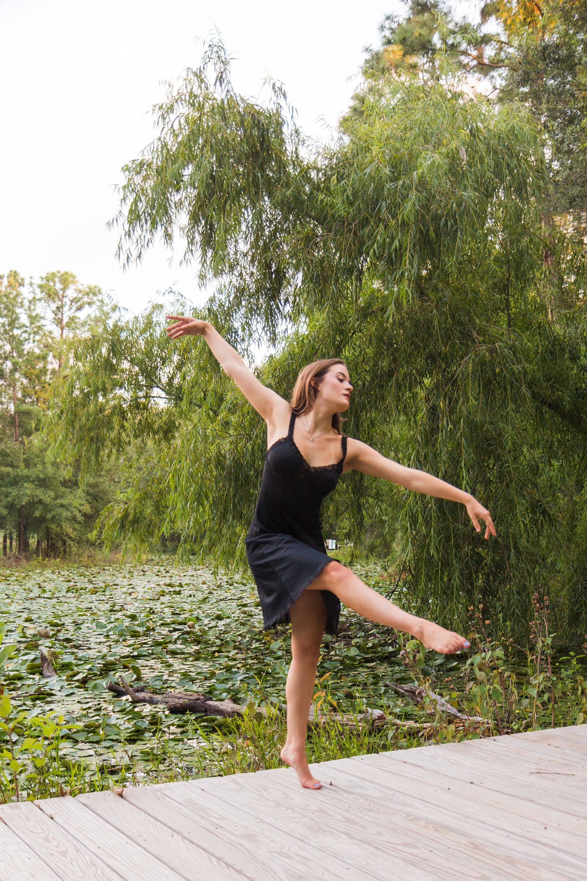A dancer in a black dress performs a pose on a wooden boardwalk next to a pond filled with lily pads and weeping willows.
