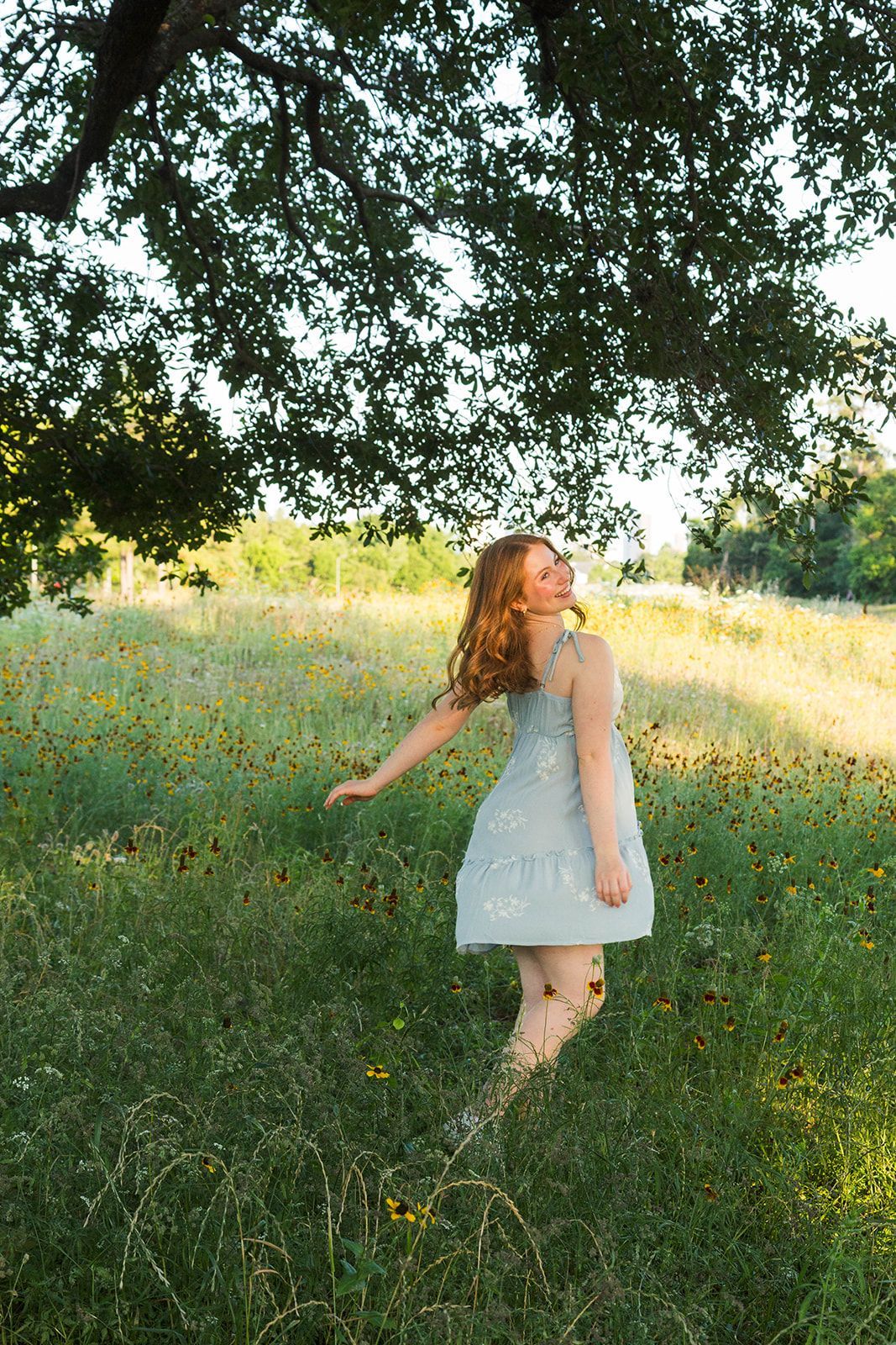 A person with long red hair wearing a light-colored dress spins in a sunlit meadow filled with tall grass and wildflowers.