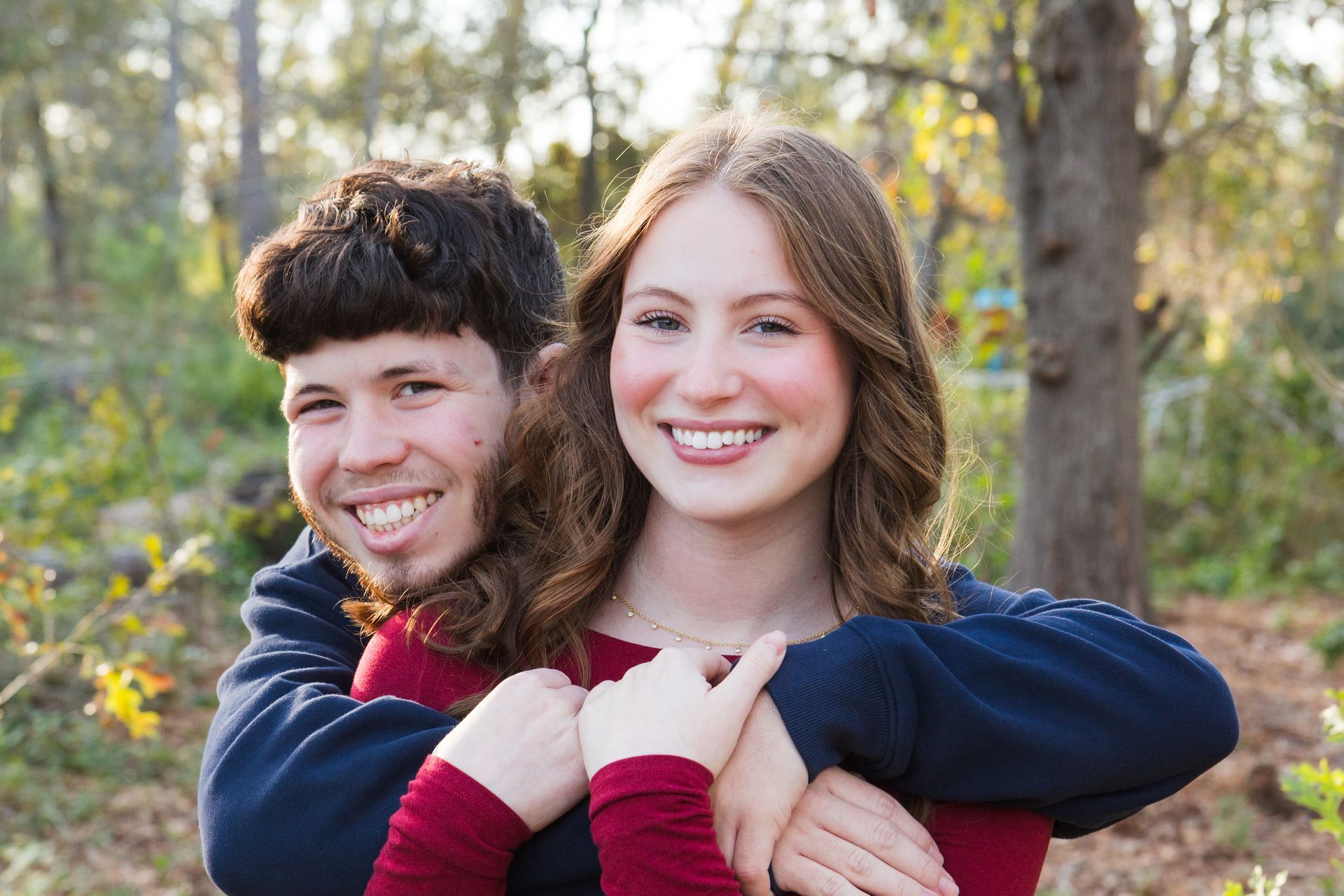 A person in a dark blue sweater hugging another person in a red top from behind, both smiling outdoors among trees.