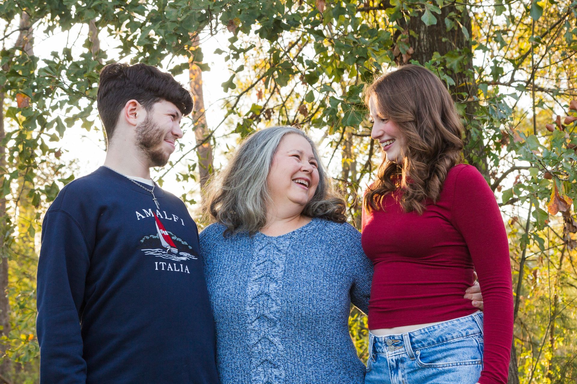 A person with gray hair stands between two younger people in a sunlit, wooded area; all three are smiling and talking.