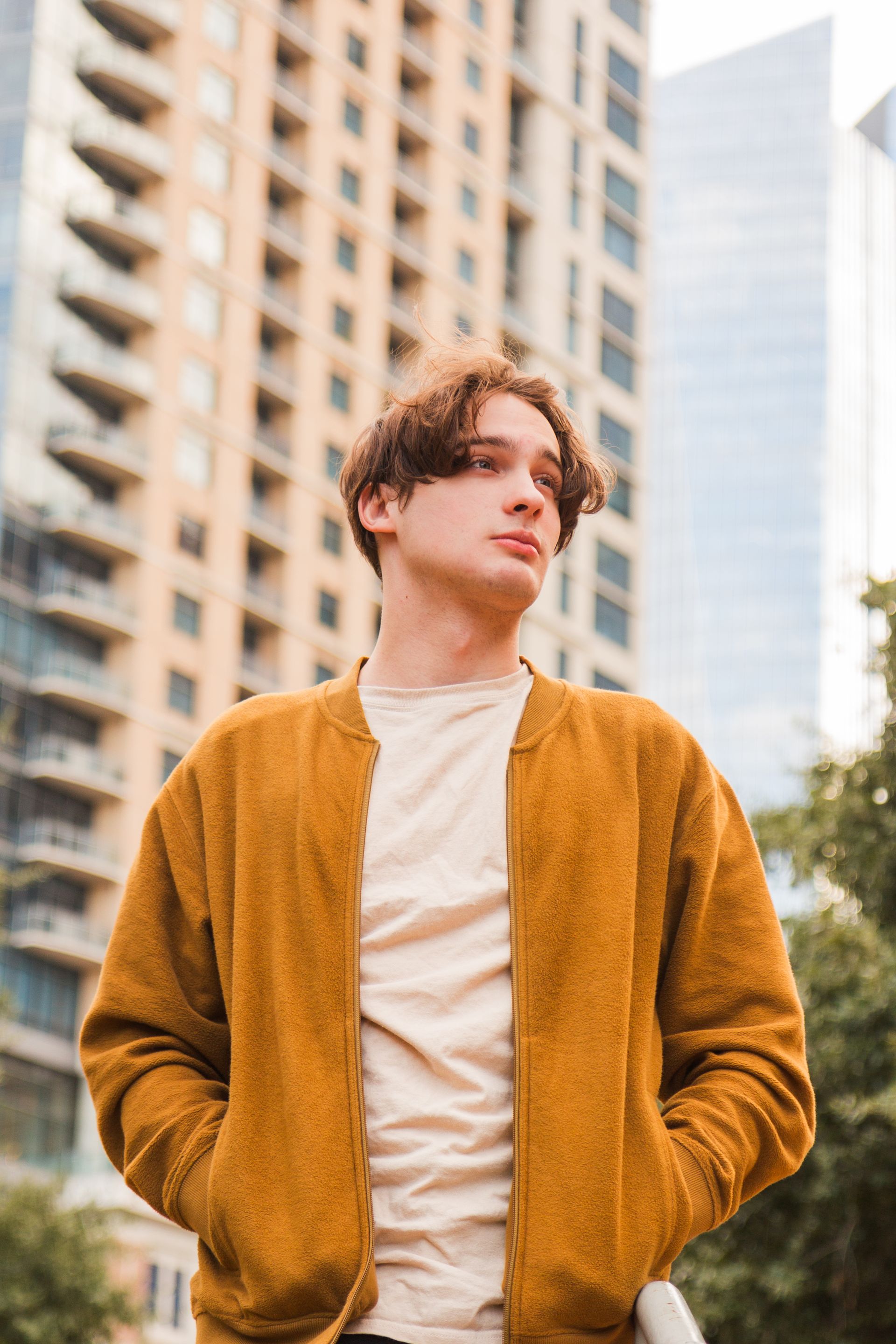 A person with wavy brown hair stands in front of tall city buildings wearing a mustard-yellow bomber jacket and beige top.