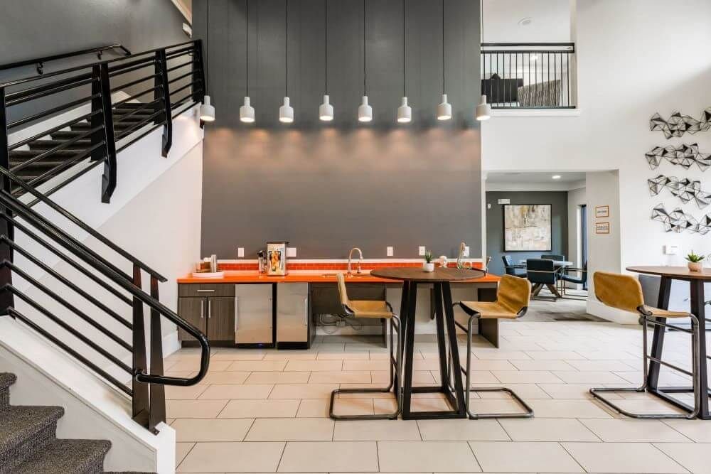Interior common area with stairs, a bar with stools, and overhead lighting against a gray wall.