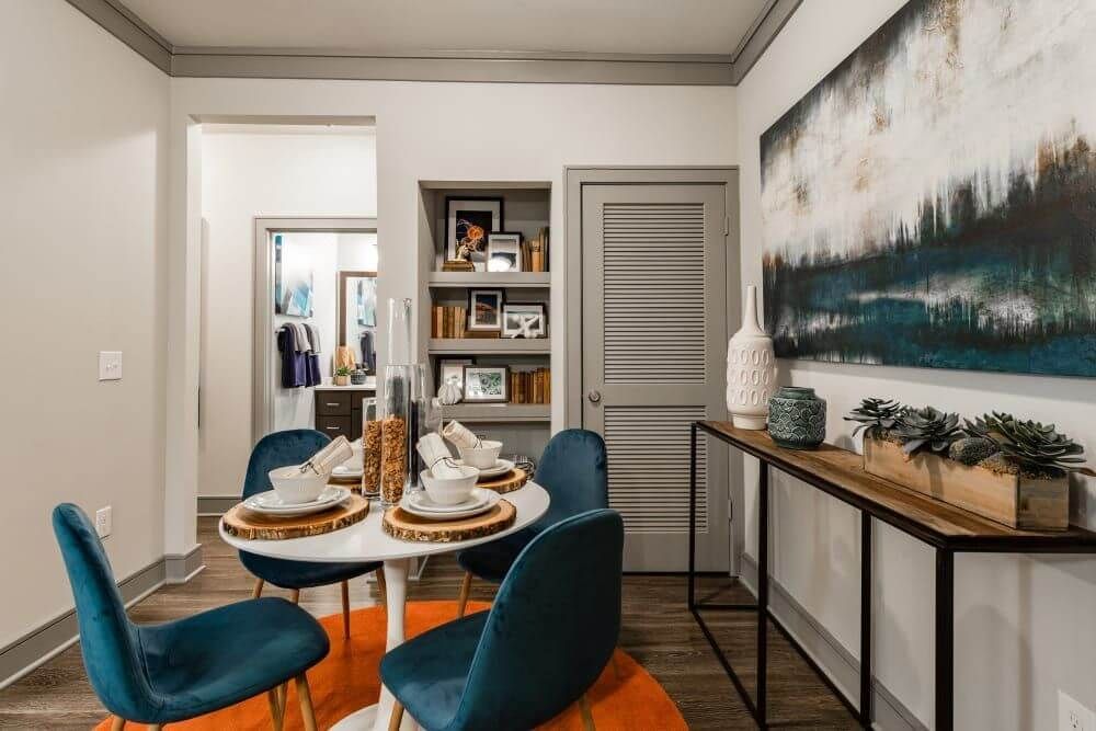 Dining area with white table, teal chairs, orange rug, and artwork.