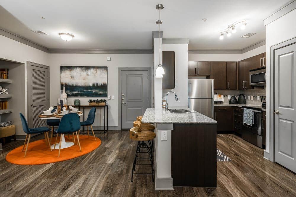 Modern kitchen and dining area with an island, dark cabinets, and wood flooring. A table is set with blue chairs.