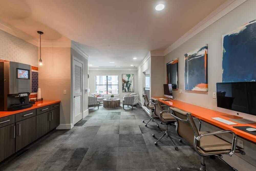 Interior of a modern office space with computer desks, coffee bar, and lounge seating. Orange and gray tones.