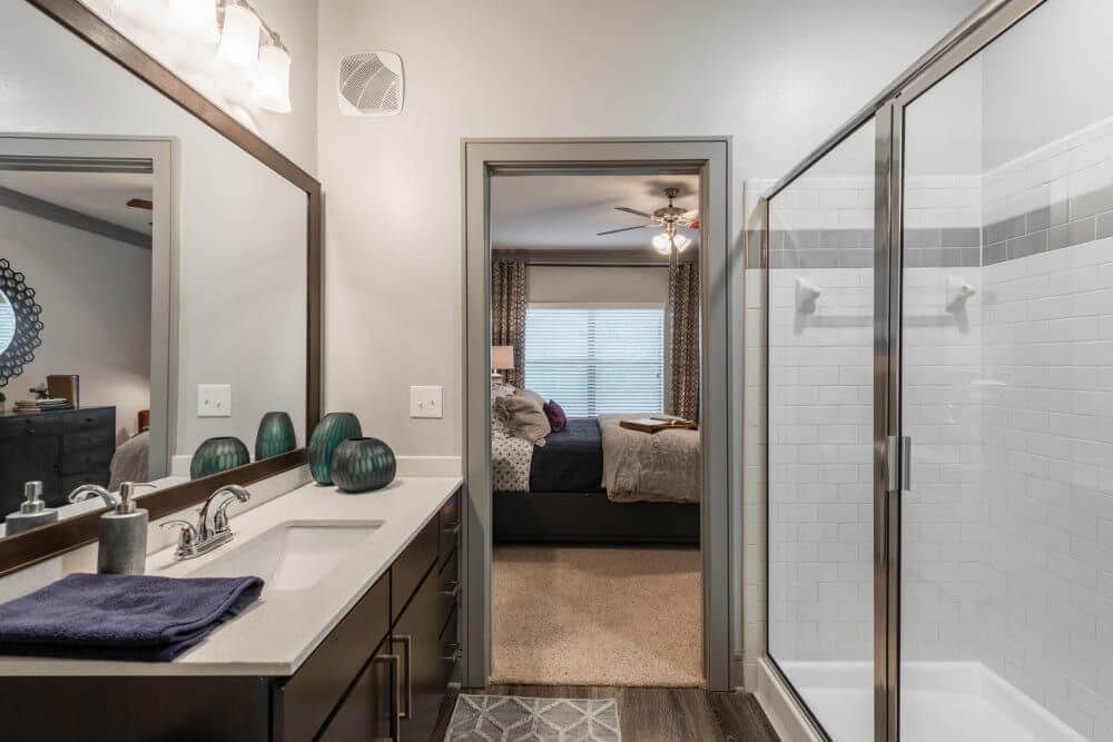 Bathroom with a vanity, shower, and doorway to a bedroom. Dark wood cabinets, white countertop, and gray walls.