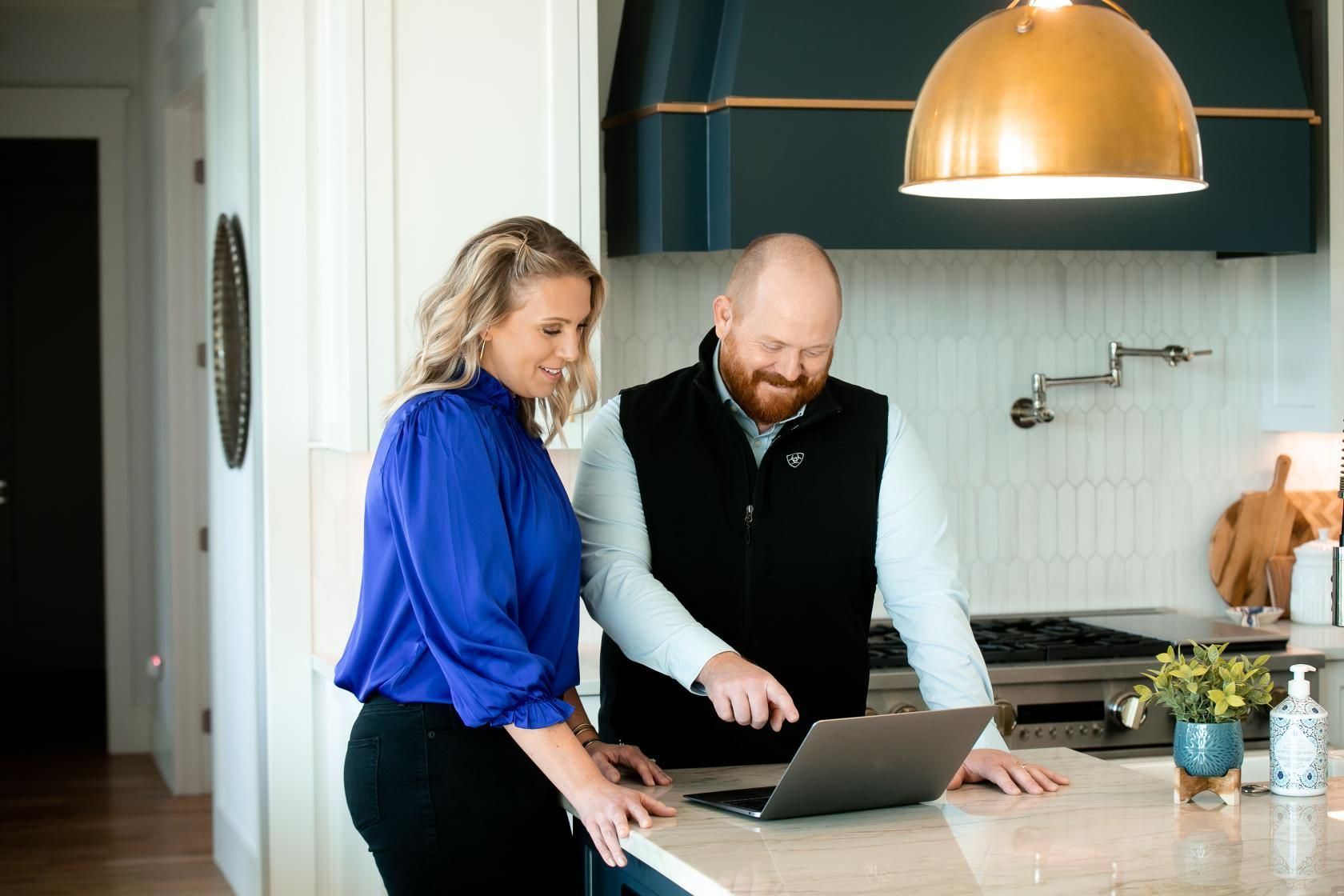 A man and a woman are standing in a kitchen looking at a laptop.