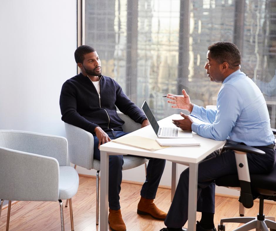 Two men are sitting at a table with a laptop and talking to each other.