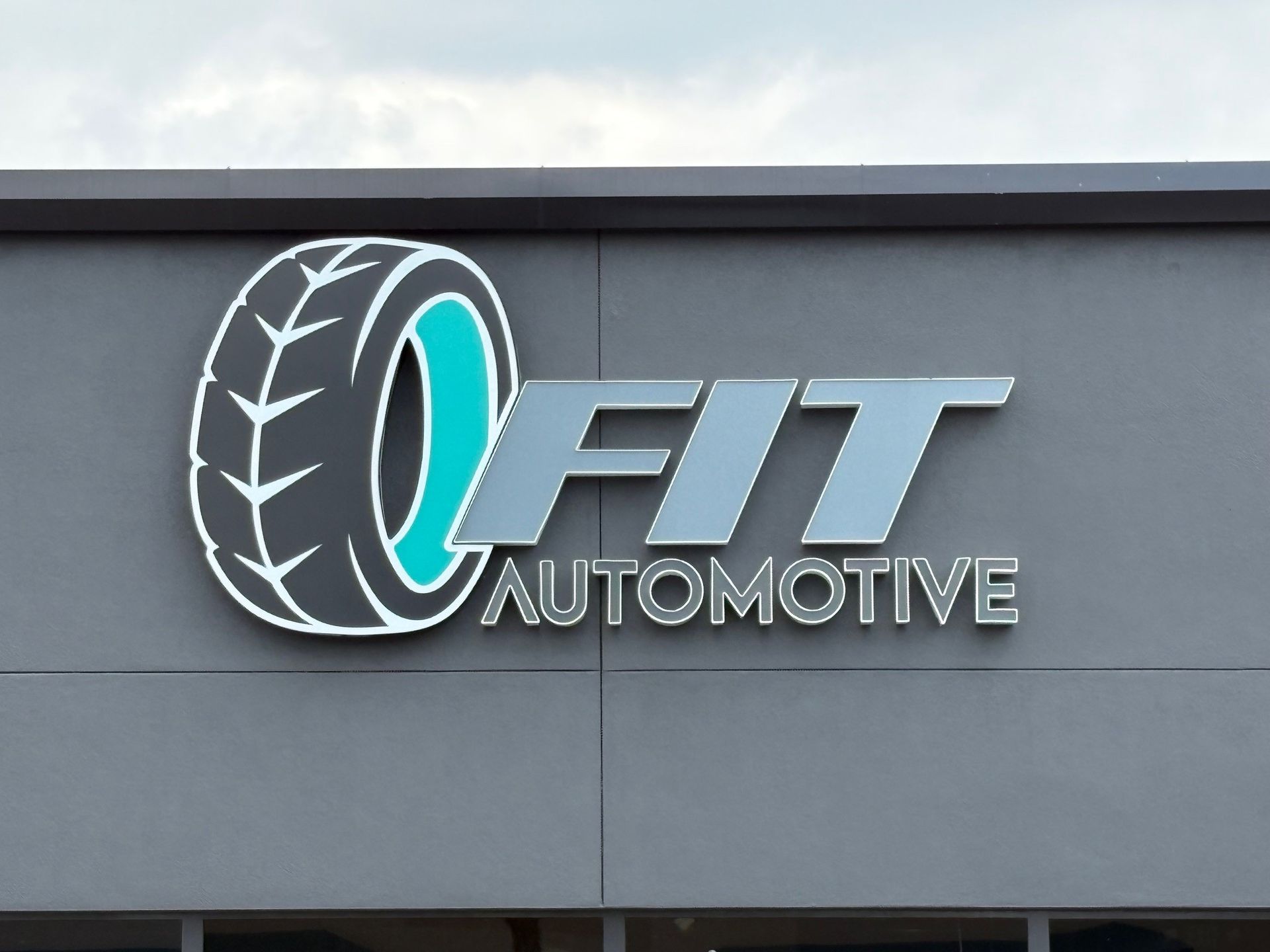 Sign for Fit Automotive, gray building, tire graphic, teal and silver text.