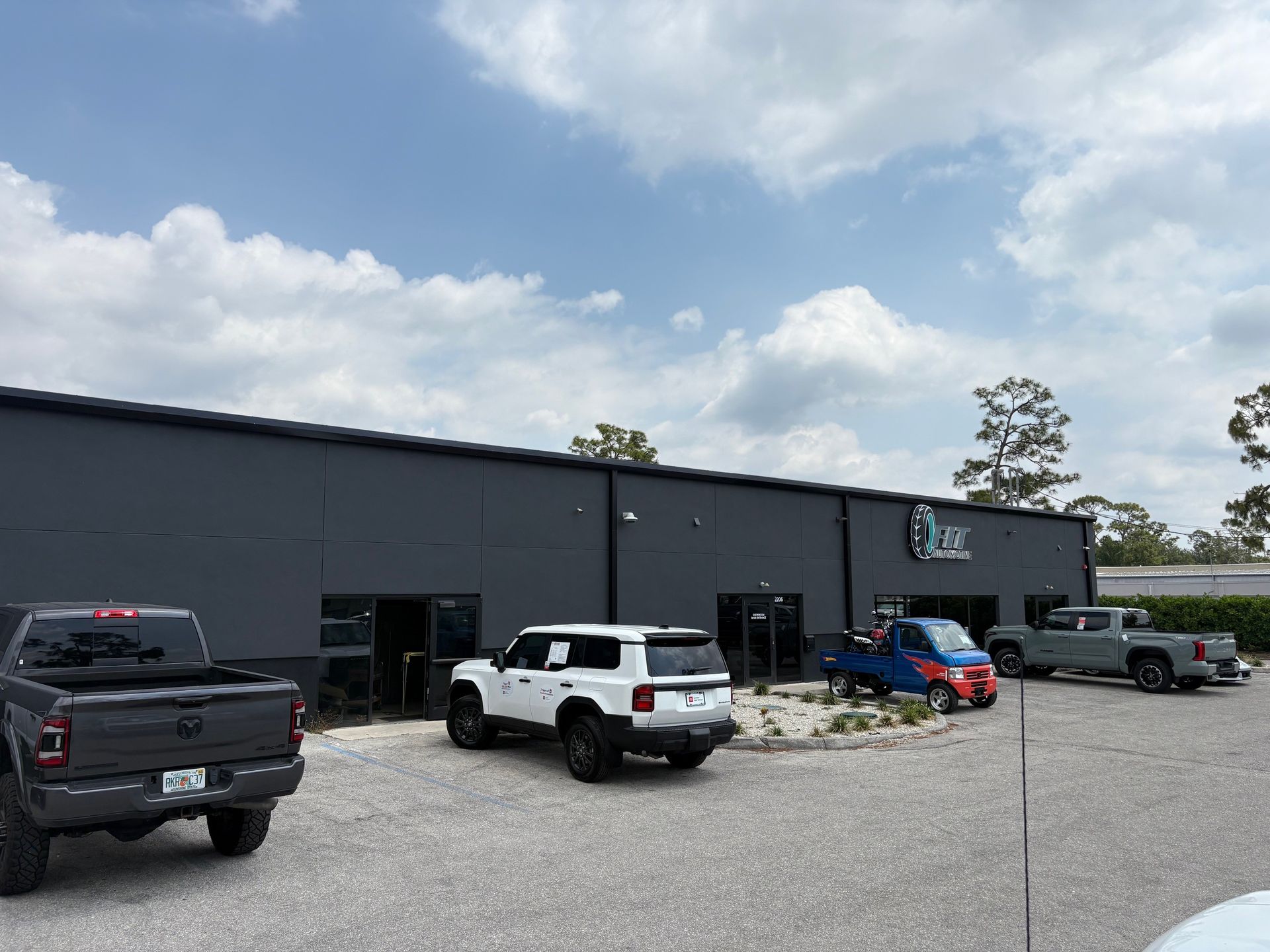 Gray building with multiple vehicles parked outside on a gravel lot under a cloudy sky.