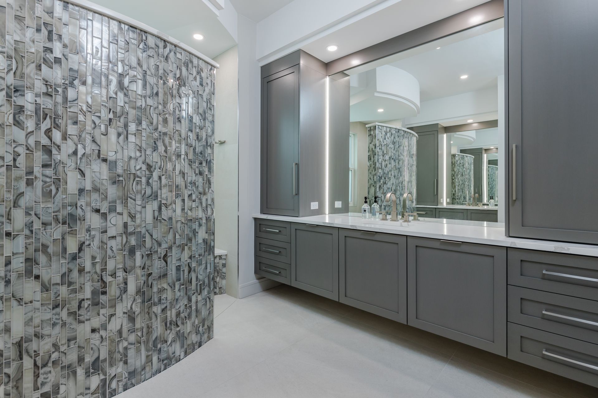 A bathroom with gray cabinets , a large mirror and a walk in shower.