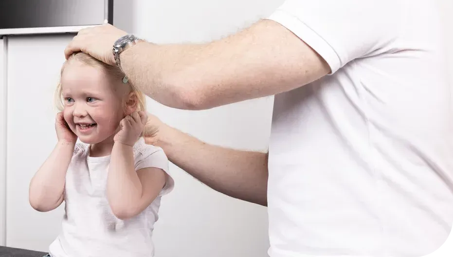 Person som berører et barns hode og ører. Barnet smiler og dekker til ørene. Hvite skjorter, innendørs.