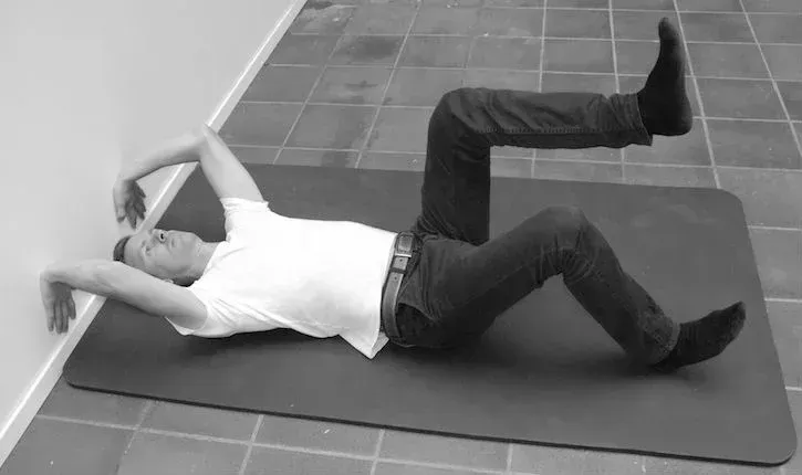 Person liggende på matte, ett ben hevet, berører veggen med armene. Innendørs setting.