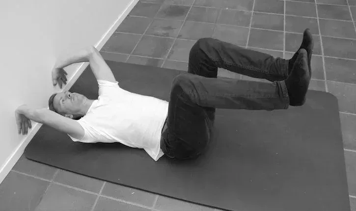 Mann ligger på ryggen på matten med beina hevet, føttene opp, armene mot veggen.