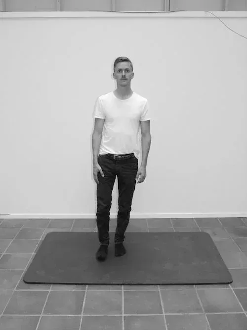 Mann i hvit t-skjorte og svarte bukser står på matte, nær en hvit vegg.