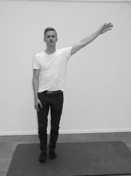 Mann i hvit t-skjorte og jeans, med arm utstrakt til siden, stående på en matte, mot en hvit vegg.