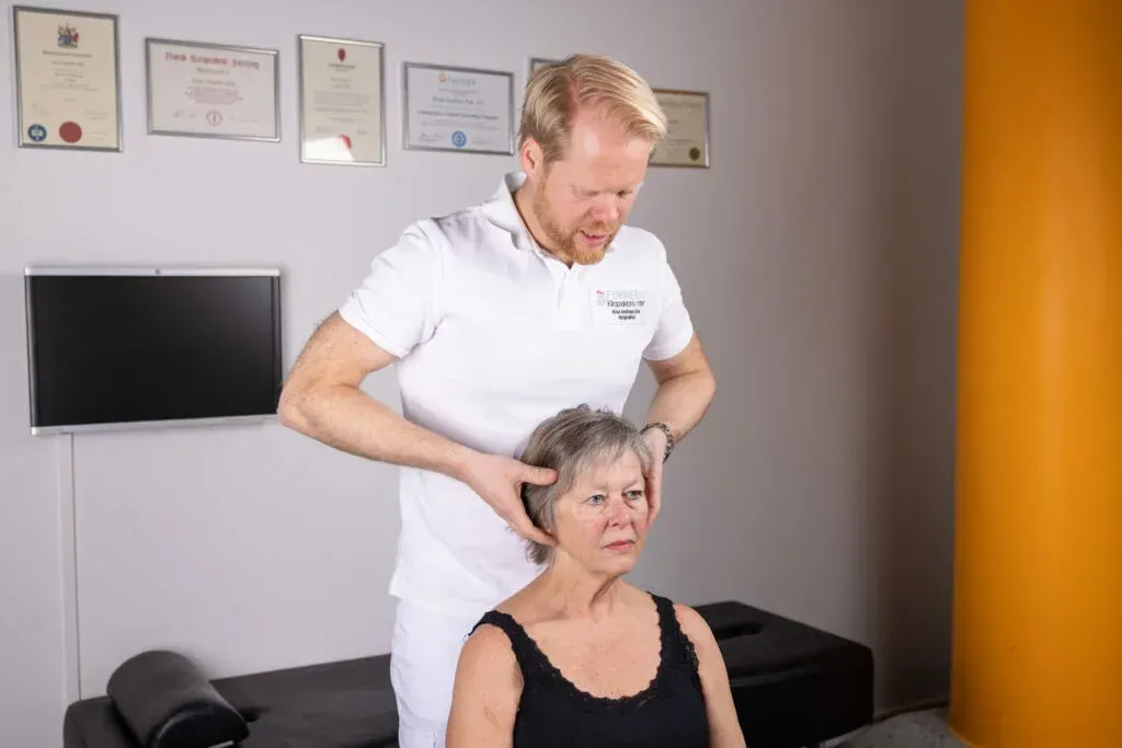 En helsepersonell undersøker en persons nakke. De er på en klinikk, i nærheten av en sofa.