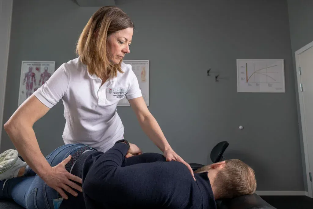 Kvinne i hvit skjorte og jeans hjelper en person på en massasjebenk med ryggsmerter. Grå vegger, to diagrammer.