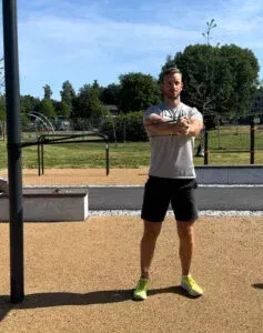 Mann står med armene i kors i parken; iført svarte shorts, grå skjorte og gule joggesko.