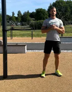 Mann trener utendørs med motstandsbånd, nøytralt uttrykk. Shorts, sportssko, park.