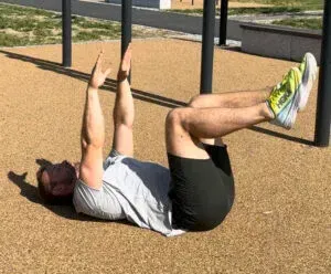 Person liggende på ryggen, armene hevet, bena bøyde, utfører en øvelse utendørs på brun overflate.