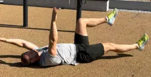 Person som ligger på bakken og trener, strekker ut høyre arm og venstre ben. Utendørs.