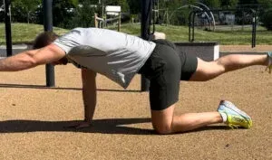 Mann i grå skjorte og svarte shorts gjør en fugl-hund-øvelse på en utendørs overflate.