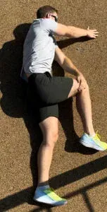 Mann i grå skjorte og svarte shorts strekker seg på bakken med utstrakt arm og bøyd ben i kneet.