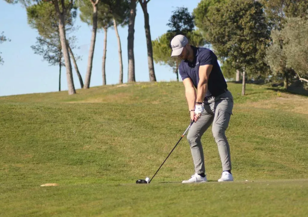 Mann i grå bukser og marineblå skjorte svinger en golfkølle på en grønn bane.