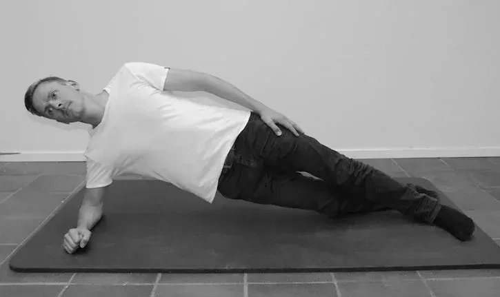 Mann i hvit skjorte og jeans utfører en sideplanke på en matte.