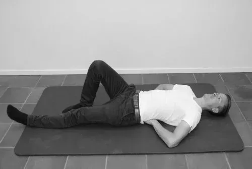 Mann ligger på ryggen, ett ben strukket ut, ett kne bøyd, hendene under korsryggen, på matten.