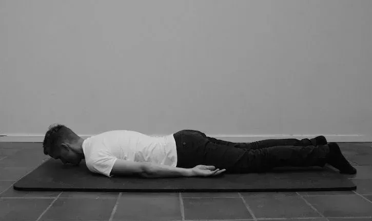 Personen ligger på mage på en matte, armene langs sidene, hodet vendt til siden og beina strukket ut.